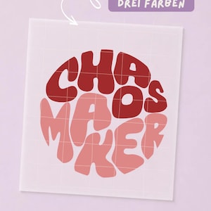 Könnte beinhalten: Ein quadratischer Vinyl-Aufkleber mit dem Text CHAOS MAKER in kreisförmiger Anordnung. Die Buchstaben sind in Dunkelrot, Hellrosa und Lachsfarben gehalten. Ein violettes rechteckiges Etikett darüber trägt die Aufschrift ERHÄLTLICH IN DREI FARBEN.