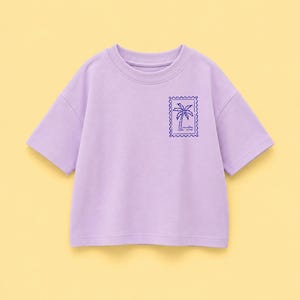 Könnte beinhalten: Ein hellviolettes T-Shirt mit kurzen Ärmeln und Rundhalsausschnitt. Auf der linken Brust befindet sich eine dunkelviolette, rechteckige Grafik, die an eine Briefmarke erinnert, mit einer Palme und dem Text vacation mode.