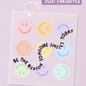 Könnte beinhalten: Ein quadratisches Blatt mit neun pastellfarbenen Smileys in Pink, Gelb, Lila, Pfirsich, Blau und Grün. Dunkellila Text verläuft geschwungen in der Mitte: "BE THE REASON SOMEONE SMILES TODAY". Ein lila Etikett oben trägt die Aufschrift "ERHÄLTLICH IN ZWEI VARIANTEN".