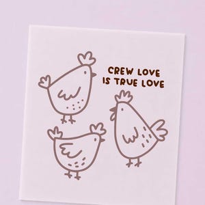 Könnte beinhalten: Eine quadratische Karte mit hellviolettem Hintergrund und drei braunen Strichzeichnungen von Hühnern. Der Text "CREW LOVE IS TRUE LOVE" ist in braunen, abgerundeten Großbuchstaben über den Hühnern auf der rechten Seite aufgedruckt.
