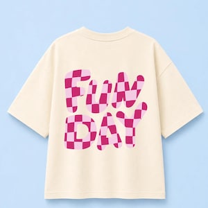 Könnte beinhalten: Ein cremefarbenes T-Shirt mit kurzen Ärmeln und dem Schriftzug "FUN DAY" in einer großen, fetten Bubble-Schriftart. Die Buchstaben sind mit einem rosa und dunkelmagenta karierten Muster gefüllt, das einen Kontrast zum hellen Stoff bildet.