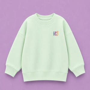 Könnte beinhalten: Ein helles mintgrünes Sweatshirt mit Rundhalsausschnitt, langen Ärmeln und gerippten Bündchen. Auf der Brust befindet sich der Schriftzug HeJ, gestickt in kleinen, bunten Buchstaben mit einem winzigen orangefarbenen Herz über dem j.