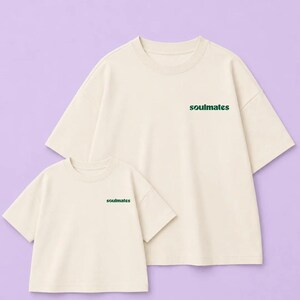 Könnte beinhalten: Zwei cremefarbene T-Shirts mit kurzen Ärmeln und Rundhalsausschnitt. Jedes Shirt ist auf der linken Brustseite mit dem Wort "soulmates" in einer dunkelgrünen, abgerundeten Kleinschrift bedruckt. Die T-Shirts sind in verschiedenen Größen.