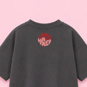Könnte beinhalten: Ein dunkel anthrazitfarbenes T-Shirt mit kurzen Ärmeln und einer kreisförmigen Grafik auf der oberen Brust. Die Grafik enthält den Text "CHAOS MAKER" in einer stilisierten, blasenartigen Schrift, wobei "CHAOS" dunkelrot und "MAKER" hellrosa ist.