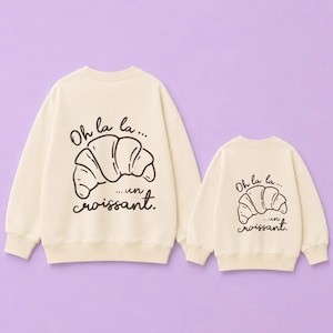 Può includere: Due felpe girocollo color crema con un'illustrazione a linee marrone scuro di un croissant. Sopra il croissant c'è il testo "Oh la la..." e sotto il testo "...un croissant." Le felpe presentano un design identico.