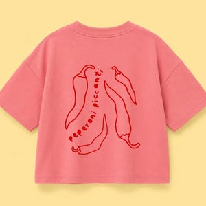 以下が含まれることがあります： 4本の唐辛子の赤いグラフィックプリントと、その間に縦書きで「peperoni piccanti」という文字がデザインされた、半袖のピンク色のTシャツ。クルーネックで、ゆったりとしたボックス型のシルエットが特徴です。