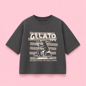 Könnte beinhalten: Ein anthrazitfarbenes Kurzarm-T-Shirt mit einer Grafik eines Hundes auf einem Roller, der eine Eistüte hält. Das Design enthält den Text "GELATO TIME" und "The best time for ice cream is always" in Weiß auf horizontalen Streifen gedruckt.