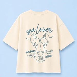Könnte beinhalten: Ein cremefarbenes T-Shirt mit kurzen Ärmeln und einer dunkelblauen Strichzeichnung eines Hummers. Der Text lautet "sea lover" oben, "ocean is calling..." links und "let's catch some lobster!" rechts neben der Hummergrafik auf dem T-Shirt.