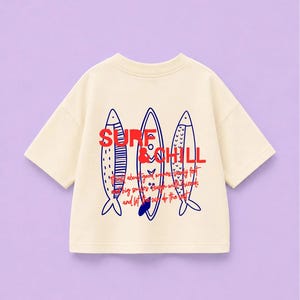 Op de afbeelding: Een crèmekleurig T-shirt met korte mouwen en een grafische print van drie gestileerde vissen en surfplanken in donkerblauw. De tekst SURF & CHILL is in het rood gedrukt, vergezeld van kleinere rode cursieve tekst.