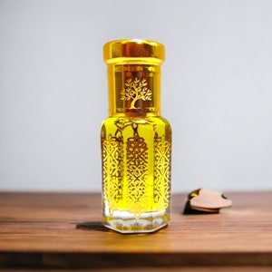 May include: A decorative perfume bottle with a gold lid and gold patterns. The bottle contains a yellow liquid and is on a wooden surface. A small, brown leather tag is next to it. The bottle's design is ornate.