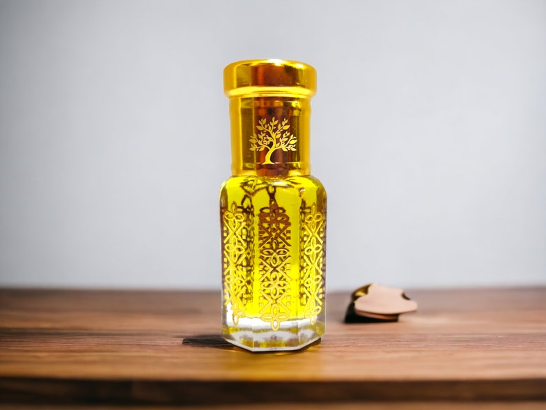 May include: A decorative perfume bottle with a gold lid and gold patterns. The bottle contains a yellow liquid and is on a wooden surface. A small, brown leather tag is next to it. The bottle's design is ornate.