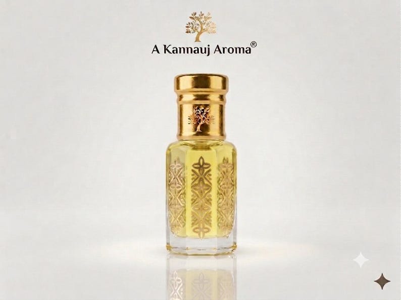 May include: A clear glass perfume bottle with a gold cap and a gold-toned geometric pattern on the sides. The bottle contains a pale yellow liquid. Above the bottle, the text A Kannauj Aroma is printed with a small tree icon above it.