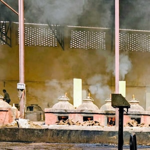 May include: A row of industrial clay ovens with conical lids, each emitting thick gray smoke into a large warehouse space. The ovens are constructed from reddish-brown masonry and are fueled by piles of wood burning at their bases.
