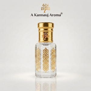 May include: A clear glass perfume bottle with a gold cap and gold decorative patterns on the sides. The text A Kannauj Aroma is printed above the bottle with a tree icon. The bottle has a hexagonal shape and a reflective surface finish.