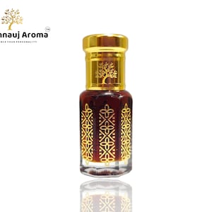 以下が含まれることがあります： 金色のキャップと複雑な金色のデザインが施された、小さな透明なガラス瓶。瓶には濃い茶色の液体が入っています。瓶には「A Kannauj Aroma」と「Enhance Your Personality」というタグラインが貼られています。