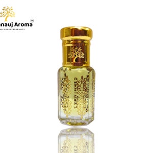 May include: A small, clear glass bottle with a gold cap and intricate gold designs. The bottle is filled with a light yellow liquid. The bottle is labelled "A Kannauj Aroma" with the tagline "Enhance Your Personality".