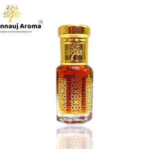 Puede incluir: Una botella dorada con un líquido marrón en su interior. La botella tiene una tapa dorada y un diseño dorado en la parte delantera. La botella está sobre una superficie blanca. El texto "A Kannauj Aroma" es visible en la etiqueta.