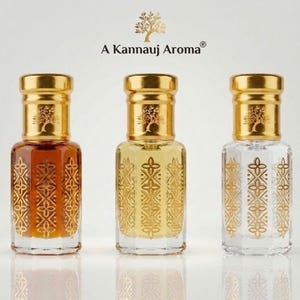 Puede incluir: Tres frascos de perfume de vidrio con tapas doradas y patrones geométricos dorados. Los frascos contienen líquidos de diferentes tonos: ámbar oscuro, amarillo claro y transparente. El texto A Kannauj Aroma aparece sobre los frascos.