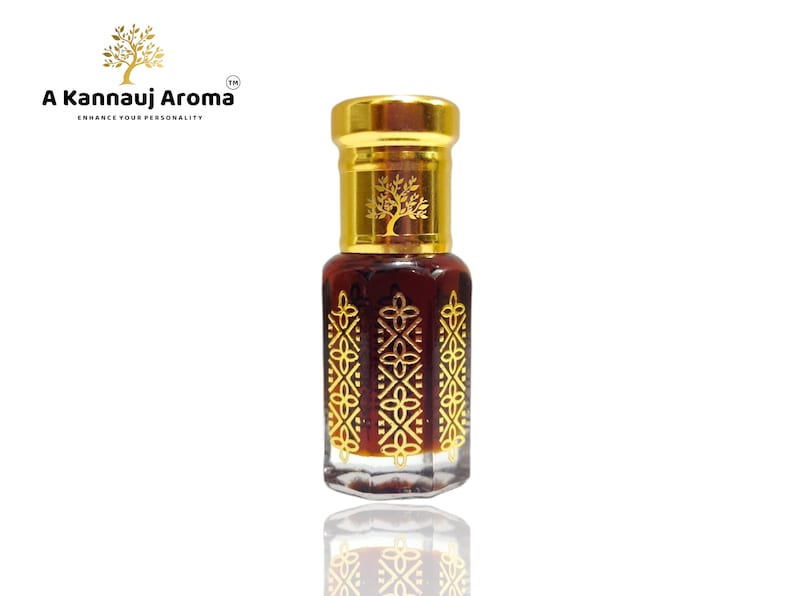 May include: A small, clear glass bottle with a gold cap and intricate gold designs. The bottle contains a dark brown liquid. The bottle is labelled "A Kannauj Aroma" with the tagline "Enhance Your Personality".