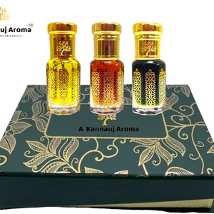 May include: A set of three small glass bottles with gold lids and decorative designs. The bottles are filled with different colored liquids and are arranged in a green box with a gold floral pattern. The box has the text "A Kannauj Aroma" on the front.