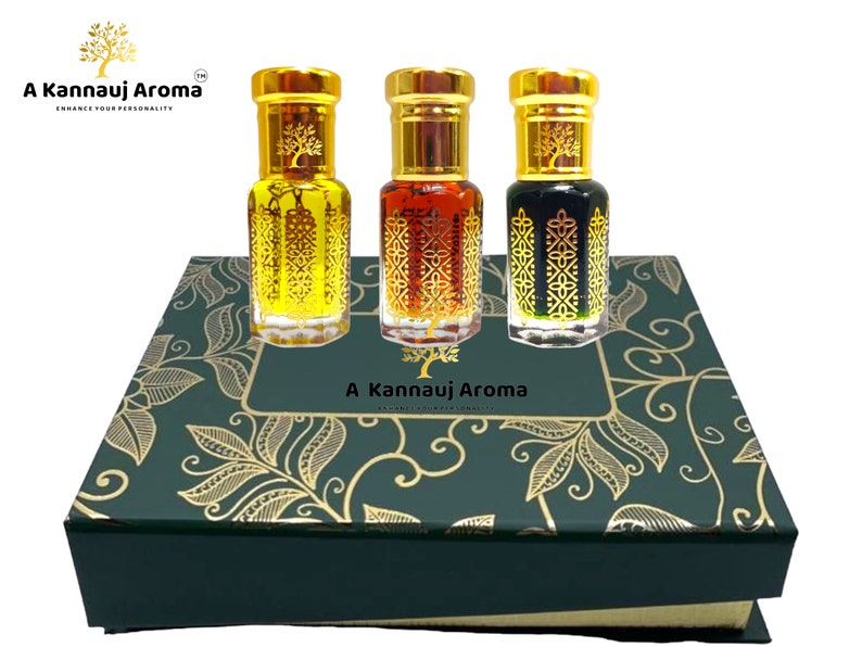 May include: A set of three small glass bottles with gold lids and decorative designs. The bottles are filled with different colored liquids and are arranged in a green box with a gold floral pattern. The box has the text "A Kannauj Aroma" on the front.