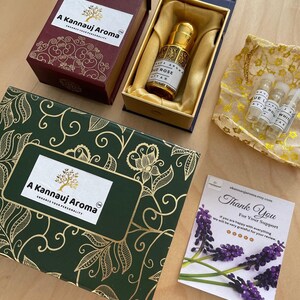 May include: A set of A Kannauj Aroma products including a large dark green box with gold floral patterns, a maroon box containing a gold-capped glass perfume bottle labeled Oud Rose Attar, three small sample vials in a gold pouch, and a thank you card.