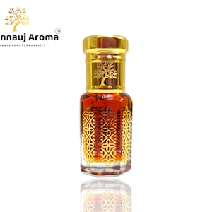 May include: A small, clear glass bottle with a gold cap and a gold design. The bottle is filled with a dark brown liquid. The bottle is labelled "A Kannauj Aroma" and "Enhance Your Personality".