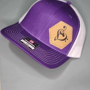 May include: Purple and white trucker hat with a cork patch featuring a musical note and heart design.
