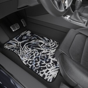 Op de afbeelding: Een zwart-witte autovloermat met luipaardprint en een aquareleffect. De mat ligt in de voetruimte aan de bestuurderskant van een auto.