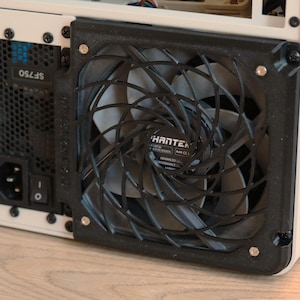May include: A computer cooling fan with a black spiral-patterned grill mounted on a white chassis. The fan features grey blades and a central Phanteks label. Adjacent to the fan is a black power supply unit labeled SF750 with a power switch.