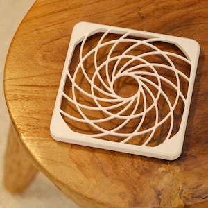 May include: A square, white coaster with a geometric, spiral design. The coaster is made of a solid material and sits on a wooden surface. The design features a series of curved lines that create a circular pattern in the center.