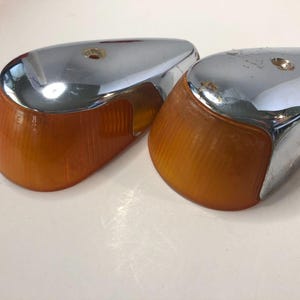 May include: A pair of automotive turn signal light assemblies featuring amber-colored, ribbed plastic lenses attached to polished chrome-finished metal housings. Each unit has a single circular mounting hole on the top surface of the chrome casing.
