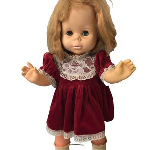May include: A doll with shoulder-length blonde hair tied with a red ribbon, featuring blue eyes and a light skin tone. The doll wears a short-sleeved, dark red velvet dress with a white lace collar and hem, paired with white shoes.