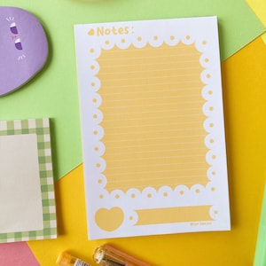 Peut inclure: Un bloc-notes ligné jaune avec une bordure blanche festonnée et un motif en forme de cœur. Le bloc-notes porte le texte "Notes :" en haut et "@tar.tea.no" en bas.