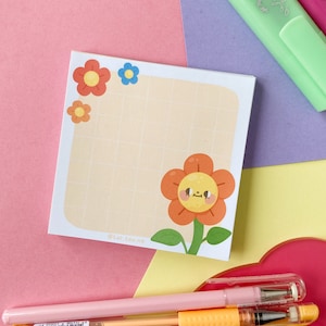 Può includere: Un blocco note giallo a griglia con un fiore arancione sorridente e fiori più piccoli di colore arancione e blu nell'angolo superiore. Il blocco note ha il testo "@tar.tea.me" nella parte inferiore.