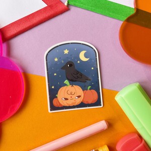 May include: A sticker featuring a black crow perched atop three orange pumpkins with a face. The background is a dark blue night sky with a crescent moon and stars. The sticker has a white border.