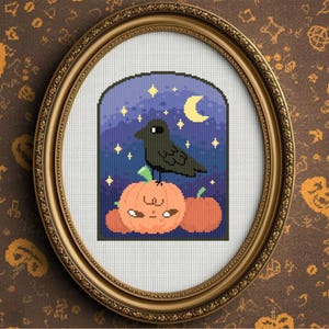 May include: An oval, gold-framed cross-stitch picture. It depicts a black crow on an orange pumpkin with a smiling face. The background shows a night sky with a crescent moon and stars. The artwork is framed.