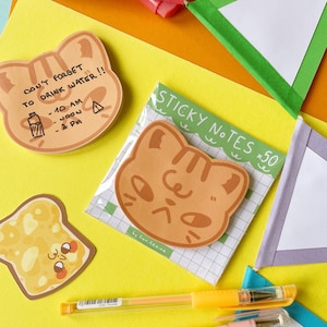 Peut inclure: Des post-it en forme de chat et une note en forme de toast sur fond jaune. Une note indique "N'OUBLIEZ PAS DE BOIRE DE L'EAU !!" avec un calendrier de rappel. Les post-it sont emballés dans un motif quadrillé vert et blanc.