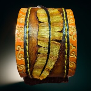Puede incluir: Un brazalete de cuero marrón con un diseño de pluma de color dorado y costuras naranjas. El brazalete tiene un patrón decorativo en los bordes.