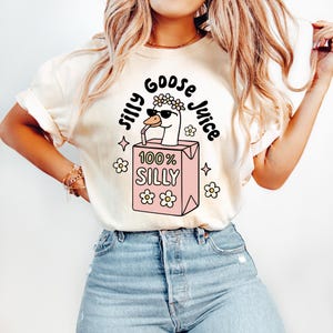 May include: Cream-colored t-shirt with a whimsical graphic. The design features a goose wearing sunglasses and a flower crown, drinking from a pink juice box labeled "100% Silly." The words "Silly Goose Juice" are arched around the goose. Floral and star accents complete the design.