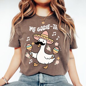 May include: Brown t-shirt with a cartoon goose wearing a sombrero and holding maracas. The text "Me Goose-Ta" is above the goose. Musical notes and flowers surround the goose, creating a festive design.