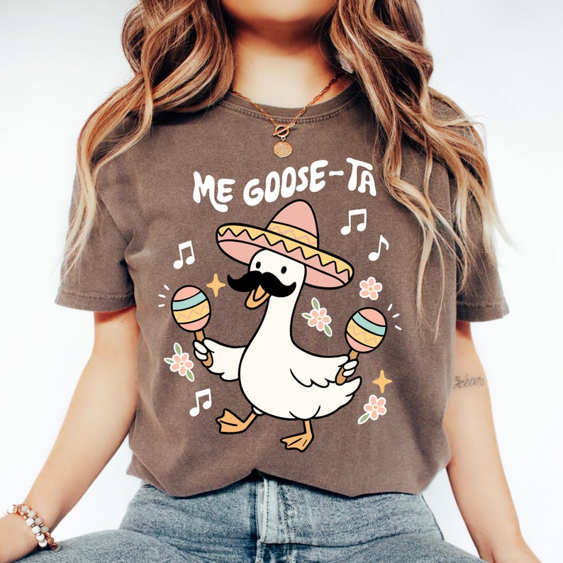 May include: Brown t-shirt with a cartoon goose wearing a sombrero and holding maracas. The text "Me Goose-Ta" is above the goose. Musical notes and flowers surround the goose, creating a festive design.