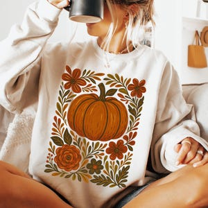 May include: Cream-colored sweatshirt featuring a large orange pumpkin design surrounded by floral and leaf accents in shades of brown, orange, and green. The design is centered on the front of the sweatshirt, creating a fall-themed aesthetic.