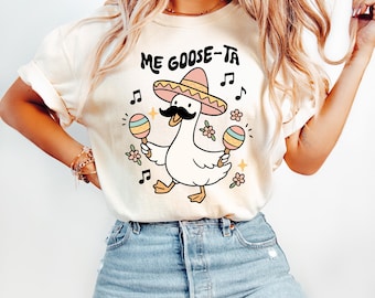 May include: Cream-colored t-shirt with the text "ME GOOSE-TA" and a cartoon goose wearing a sombrero and a mustache, holding maracas. Musical notes and floral accents surround the goose, creating a festive design.
