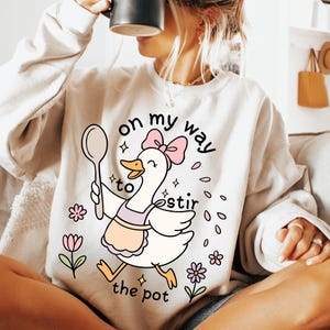 May include: Cream-colored sweatshirt with a graphic of a cartoon goose wearing an apron and a pink bow. The goose holds a spoon and the text reads "on my way to stir the pot."