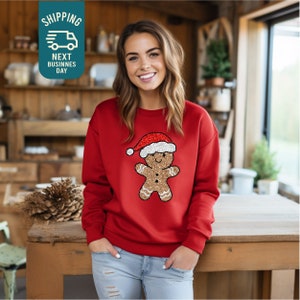 May include: Red Christmas sweatshirt with a gingerbread man wearing a Santa hat. The gingerbread man is made of glitter and is a festive design.