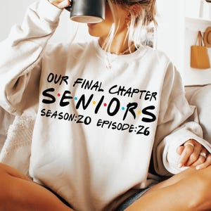 May include: Cream-colored sweatshirt with the text "OUR FINAL CHAPTER SENIORS SEASON:20 EPISODE:26" in black, red, blue, and yellow lettering. The sweatshirt is a crew neck style and is being worn by a person holding a black mug.