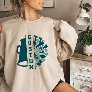 May include: A beige sweatshirt with a teal and blue cheerleader design and the word "CUSTOM" printed vertically. The design includes a megaphone and pom-poms. The sweatshirt is a long-sleeved, crew neck style.
