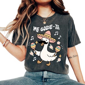 May include: Dark gray t-shirt featuring a cartoon goose wearing a sombrero and a mustache, holding maracas. The text "Me Goose-Ta" is above the goose. Musical notes and flowers surround the goose, creating a festive design.