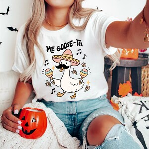 May include: White t-shirt with the text "Me Goose-Ta" and a cartoon goose wearing a sombrero and mustache, holding maracas. The design includes musical notes and flowers, creating a festive and playful vibe.